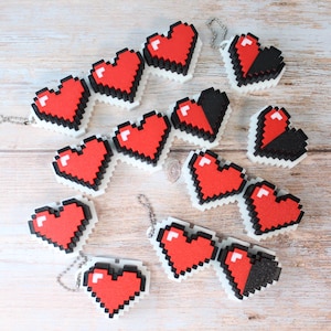 Könnte beinhalten: Pixelige herzförmige Schlüsselanhänger in Rot, Schwarz und Weiß. Die Schlüsselanhänger sind auf einer hellen Holzoberfläche angeordnet. Die Herzen sind im Retro-8-Bit-Stil gehalten, mit weißem Rand und silberner Kette.