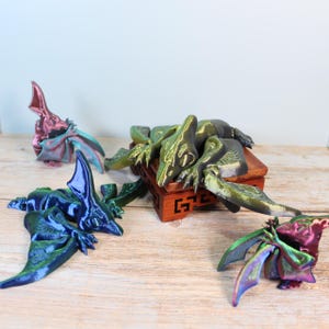 May include: Four iridescent, 3D printed pterodactyl figurines. One is green and gold, one is blue and green, one is pink and purple, and one is red and purple. The green and gold pterodactyl is lying on a small wooden stand.