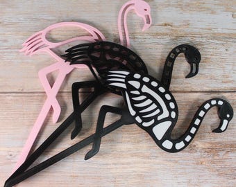 May include: Three flamingo-shaped decorative items. Two are pink, and one is black with a white skeleton design. The flamingos are made of a flat material and have long legs and necks. The background is a light wood grain.