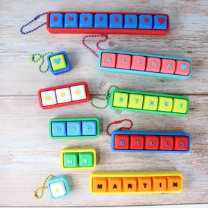 Op de afbeelding: Een verzameling kleurrijke, gepersonaliseerde naam sleutelhangers. Elke sleutelhanger toont een naam, zoals "BUFFALO" of "ALINA", gespeld op een reeks gekleurde knoppen. De sleutelhangers zijn verkrijgbaar in verschillende kleuren, waaronder rood, blauw, groen en geel.