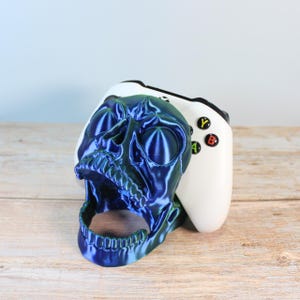Op de afbeelding: Een blauwe en groene, iriserende schedelvormige controllerstandaard met een witte Xbox-controller in de mond van de schedel.