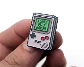 May include: A white and gray enamel pin shaped like a retro handheld video game console with a green screen that reads "I DON'T PLAY GAMES".