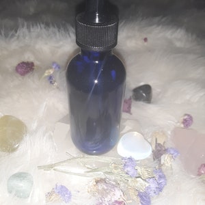 May include: A blue glass bottle with a black dropper top, surrounded by crystals and dried flowers. The bottle is sitting on a white fluffy surface.