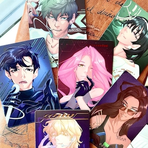 May include: A collection of anime-style character art cards. Each card features a different character with unique hairstyles, clothing, and expressions. The cards have a glossy finish and are set against a brown leather surface.