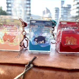May include: Three colorful milk carton-shaped keychains with cartoon characters and text. The first keychain is peach with the text "Sylvie 100% organic". The second keychain is blue with the text "Feli 100% kitty friendly". The third keychain is red with the text "Ferdie 100% AEGIR".