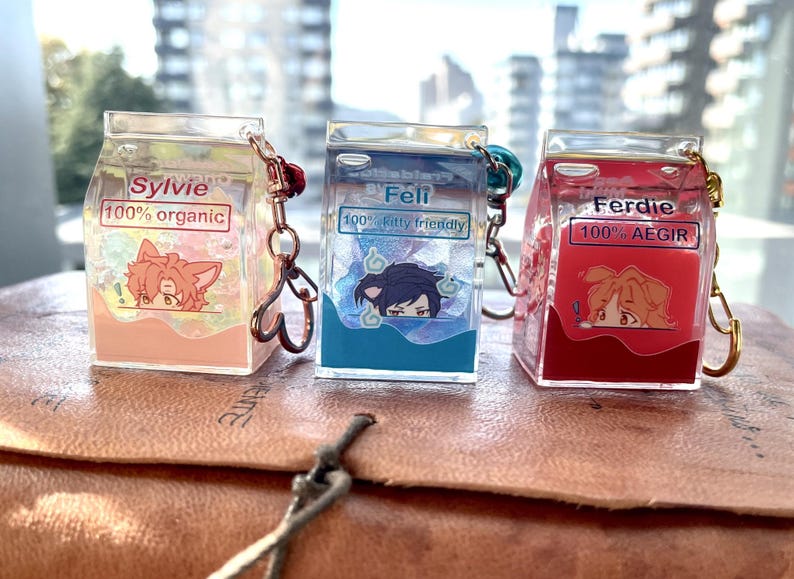May include: Three colorful milk carton-shaped keychains with cartoon characters and text. The first keychain is peach with the text "Sylvie 100% organic". The second keychain is blue with the text "Feli 100% kitty friendly". The third keychain is red with the text "Ferdie 100% AEGIR".