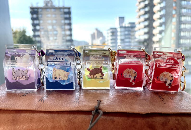 May include: Five clear plastic keychain charms shaped like milk cartons. Each charm features a different anime character with a different colored liquid inside. The text on the charms reads "Y", "Dimi 100% royal nectar", "aweno 100% NOTUS", "100% authentic", and "Edie 100% authentic".