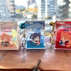 May include: Three clear plastic keychain charms shaped like milk cartons. Each charm features a different anime character and text. The first charm says "Gautier Chews" and features a pink fox character. The second charm says "Fraldarius Chews" and features a black cat character. The third charm says "Aegir Milk!" and features a red haired character.