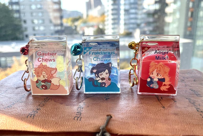 May include: Three clear plastic keychain charms shaped like milk cartons. Each charm features a different anime character and text. The first charm says "Gautier Chews" and features a pink fox character. The second charm says "Fraldarius Chews" and features a black cat character. The third charm says "Aegir Milk!" and features a red haired character.