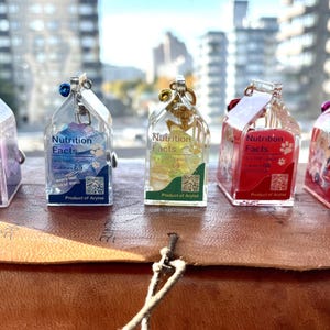 May include: Five miniature milk carton charms with different colored liquids and glitter inside. Each charm has a label that reads "Nutrition Facts" and a QR code. The charms are made of clear resin and are hanging from a silver chain.