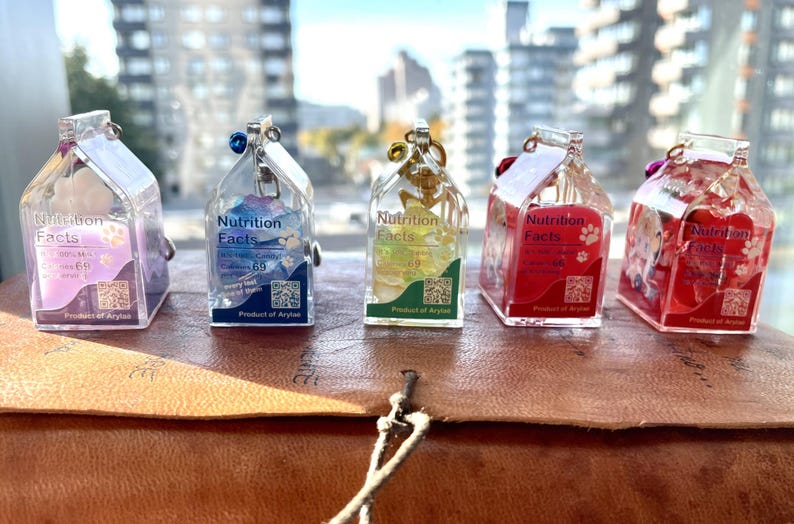 May include: Five miniature milk carton charms with different colored liquids and glitter inside. Each charm has a label that reads "Nutrition Facts" and a QR code. The charms are made of clear resin and are hanging from a silver chain.
