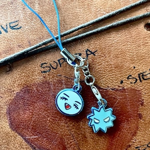 Pode incluir: Dois pequenos amuletos azul claro com detalhes em metal prateado. Um é um rosto sorridente com cílios pretos e boca vermelha. O outro tem formato de estrela com olhos brancos. Um cordão azul está preso.