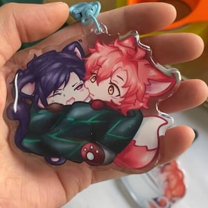 Könnte beinhalten: Ein durchsichtiger Acryl-Schlüsselanhänger mit zwei Comicfiguren. Die Figur links ist eine schwarze Katze mit lila Haaren und grüner Kleidung. Die Figur rechts ist ein roter Fuchs mit rosa Haaren und einem weißen Schwanz. Die Figuren umarmen sich.