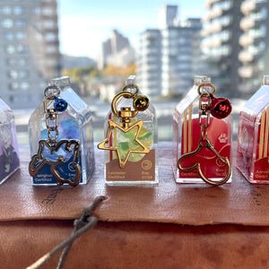 May include: Five clear plastic charm containers with gold metal charms and a gold chain. The charms are a purple paw print, a blue bird, a green star, a red heart, and a gold paw print. Each container has a label with the text "Eco Certified" and "Lancaster Gardens".