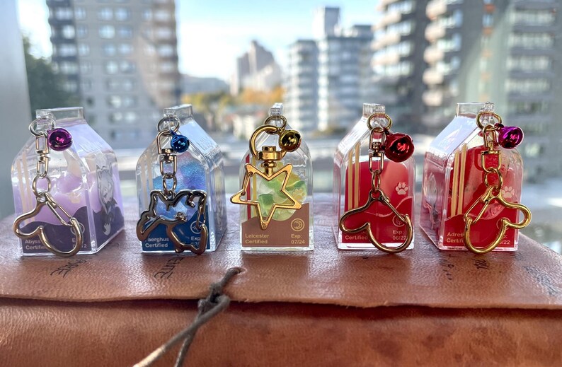 May include: Five clear plastic charm containers with gold metal charms and a gold chain. The charms are a purple paw print, a blue bird, a green star, a red heart, and a gold paw print. Each container has a label with the text "Eco Certified" and "Lancaster Gardens".