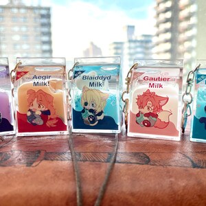 May include: Five clear plastic keychain charms shaped like milk cartons with different colored liquid inside. Each carton features a cartoon character with a different colored outfit and hair. The characters are labeled "Vestra Milk", "Aegir Milk!", "Blaiddyd Milk", "Gautier Milk", and "Fraldarius Milk".