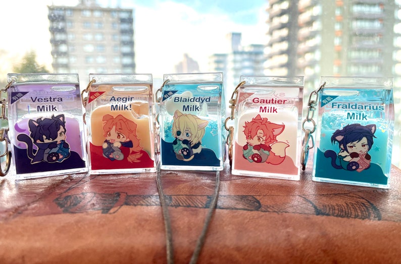 May include: Five clear plastic keychain charms shaped like milk cartons with different colored liquid inside. Each carton features a cartoon character with a different colored outfit and hair. The characters are labeled "Vestra Milk", "Aegir Milk!", "Blaiddyd Milk", "Gautier Milk", and "Fraldarius Milk".