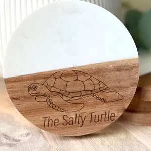 May include: A round wooden coaster with a white marble top. The coaster has a line drawing of a sea turtle and the text "The Salty Turtle".