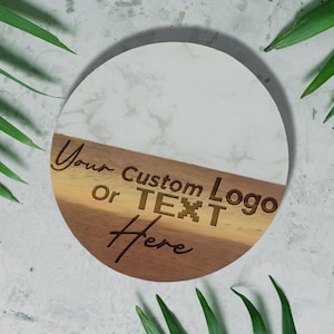 May include: A round coaster with a marble-like white top and a wooden bottom. The wooden section is divided into two tones and features the engraved text: "Your Custom Logo Or TEXT Here."