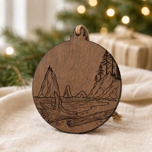 May include: Wooden Christmas ornament with a laser-etched coastal scene. The round ornament features a detailed depiction of a beach with rock formations, trees, and driftwood. It has a natural wood grain finish and a jute string for hanging.
