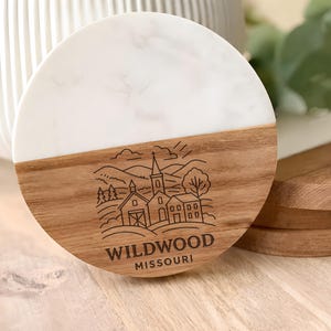 A wooden coaster with a white marble design featuring a rural landscape and a church, along with the text "WILDWOOD MISSOURI".