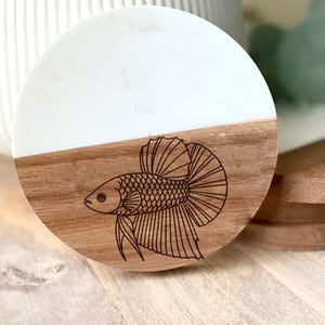 May include: A round coaster with a two-tone design. The top half is white, and the bottom half is wood-toned. A detailed black illustration of a betta fish is etched into the wood. The coaster is likely used for drinks.