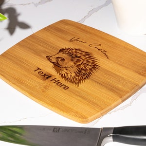 May include: A light brown square-shaped bamboo cutting board with a laser-engraved hedgehog design. The text "Your Custom Text Here" is engraved on the board.