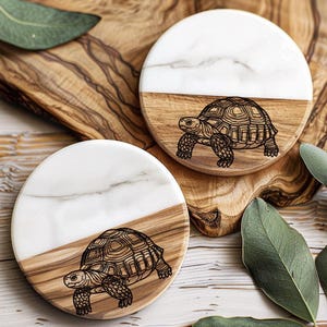 May include: Two round coasters with a tortoise design. Each coaster is half white marble and half wood. The tortoise is etched into the wood. The coasters are on a wooden surface with green leaves.