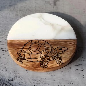 May include: A round wooden coaster with a laser-etched turtle design. The top half is white with a marble effect, and the bottom half is natural wood. The coaster is approximately 4 inches in diameter.