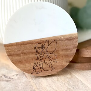May include: A round coaster with a white top and a wooden bottom. The wooden portion features a detailed engraving of a fairy. The fairy is sitting with her knees bent and her eyes closed. The coaster is approximately 4 inches in diameter.