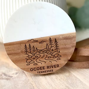 May include: A round coaster with a white top and a wood-grain bottom. The bottom features an etched design of a river scene with trees and the text "OCOEE RIVER TENNESSEE".