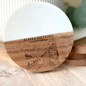 May include: A round coaster with a white top and a wood-grain bottom. The bottom features an engraved design of a road leading to houses, mountains, and a setting sun. The text "ALASKA BAYFRONT SUNSET DRIVE" is above the design.