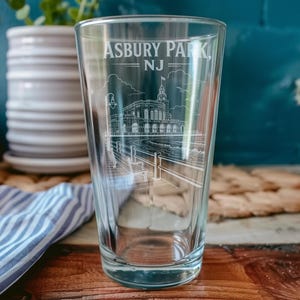 Puede incluir: Un vaso de pinta de vidrio transparente con un diseño grabado en blanco de la pasarela y el edificio del casino de Asbury Park, NJ. El texto Asbury Park, NJ está impreso en blanco en la parte superior del vaso, sobre la ilustración lineal detallada.