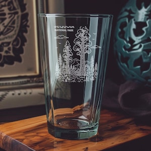 Puede incluir: Vaso de pinta transparente con un diseño blanco grabado del Parque Nacional de las Secuoyas. El diseño incluye árboles, montañas y el texto "Sequoia National Park". El vaso está sobre una superficie de madera.