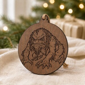 May include: A round, wooden Christmas ornament featuring a detailed engraving of a creature with a stern expression and long beard. The ornament has a natural wood grain finish and a jute string for hanging. The creature is a monster.