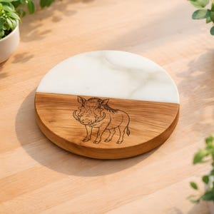 May include: A round wooden and white marble serving board. The wooden section features a detailed engraving of a warthog. The board is split in half with the top half being white marble. The board is on a light brown wooden surface.