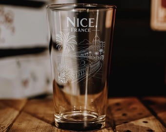 May include: A clear glass pint tumbler featuring a white etched illustration of the Nice, France coastline. The design includes a palm tree, a seaside promenade with railings, and city buildings. The text Nice, France is etched at the top.