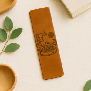 May include: A brown leather bookmark with a detailed etched illustration of a pizza. The design includes a pizza on a table, chairs, a framed picture, and a clock. The bookmark is rectangular and has rounded corners.