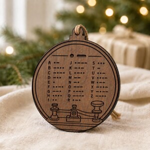May include: A round, wooden Christmas ornament with a Morse code design. The ornament is brown with engraved letters and symbols. It has a jute string for hanging and a decorative telegraph key design at the bottom.