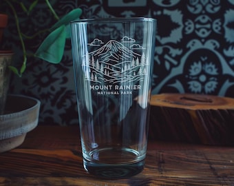 May include: Clear glass pint glass with a white graphic of Mount Rainier National Park. The design includes a mountain, trees, and the text "Mount Rainier National Park". The glass is on a wooden surface.
