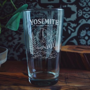 Puede incluir: Un vaso de pinta de vidrio transparente con un gráfico grabado en blanco del Parque Nacional Yosemite, CA. El diseño incluye un paisaje montañoso detallado con pinos, un río y el texto YOSEMITE NATIONAL PARK, CA impreso en la parte superior.