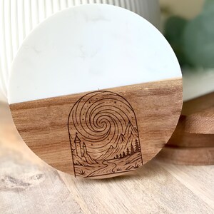 May include: Round coaster with a white top and a wooden base. The wooden base features an etched design of a mountain landscape with a castle, river, and swirling sky. The coaster is a decorative item.