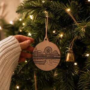 A hand is holding a wooden ornament with a drawing of a ferris wheel on a Christmas tree.