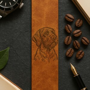 May include: A brown leather bookmark featuring an engraved portrait of a dog. The bookmark is rectangular and has a textured surface. Coffee beans, a pen, and a watch are also visible.
