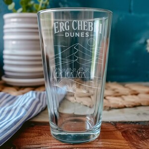 May include: A clear glass pint tumbler featuring an etched design of the Erg Chebbi dunes, a line of three camels, and the text "ERG CHEBBI DUNES" in a clean, sans-serif font. The illustration includes a sun and desert vegetation.