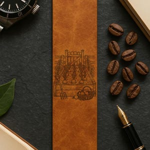 May include: A brown leather bookmark with a detailed engraved garden scene. The bookmark features a wooden fence, vegetables, and a basket of produce. The bookmark is surrounded by coffee beans, a watch, a pen, and a book.