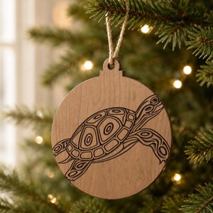 May include: A round wooden Christmas ornament with a laser-cut sea turtle design. The ornament is light brown and hangs from a natural twine string. The turtle design is detailed with etched lines.