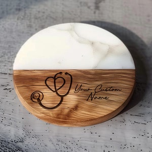 May include: A round wooden coaster with a white marble top. The coaster has a stethoscope and heart design engraved on the wood with the text "Your Custom Name".