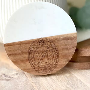 May include: A round coaster with a white top half and a wooden bottom half. The wooden portion features a compass design. The coaster is made of wood and resin. The coaster is approximately 4 inches in diameter.
