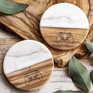 May include: Two round coasters with a gaming theme. Each coaster is half white marble and half wood, with a black outline of a game controller etched on the wood. The coasters are on a wooden surface with green leaves.